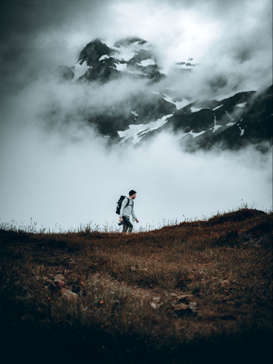 Lone hiker crossing an open ridge below a snow-streaked mountain wrapped in cloud