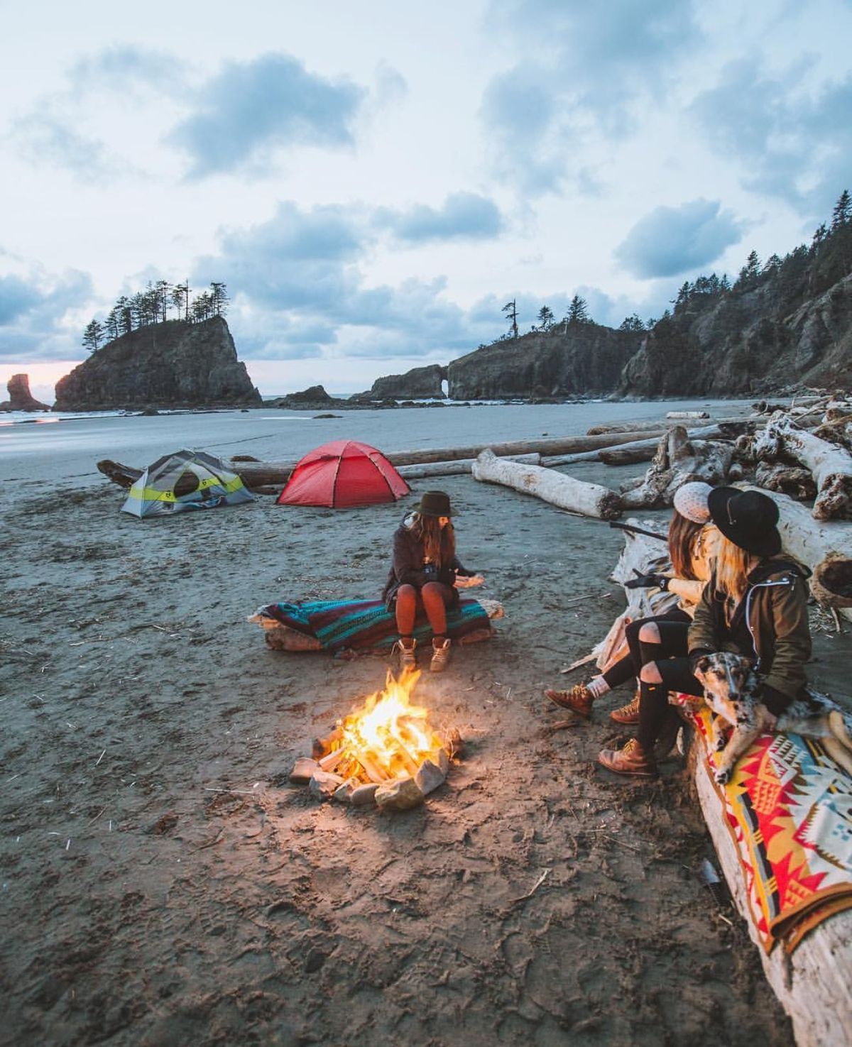 Campers around a driftwood fire on a sea-stack beach at dusk, with tents pitched on the sand behind them