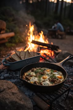 Cast-iron pan of baked eggs beside a tray of bread, cooking next to a campfire at dusk